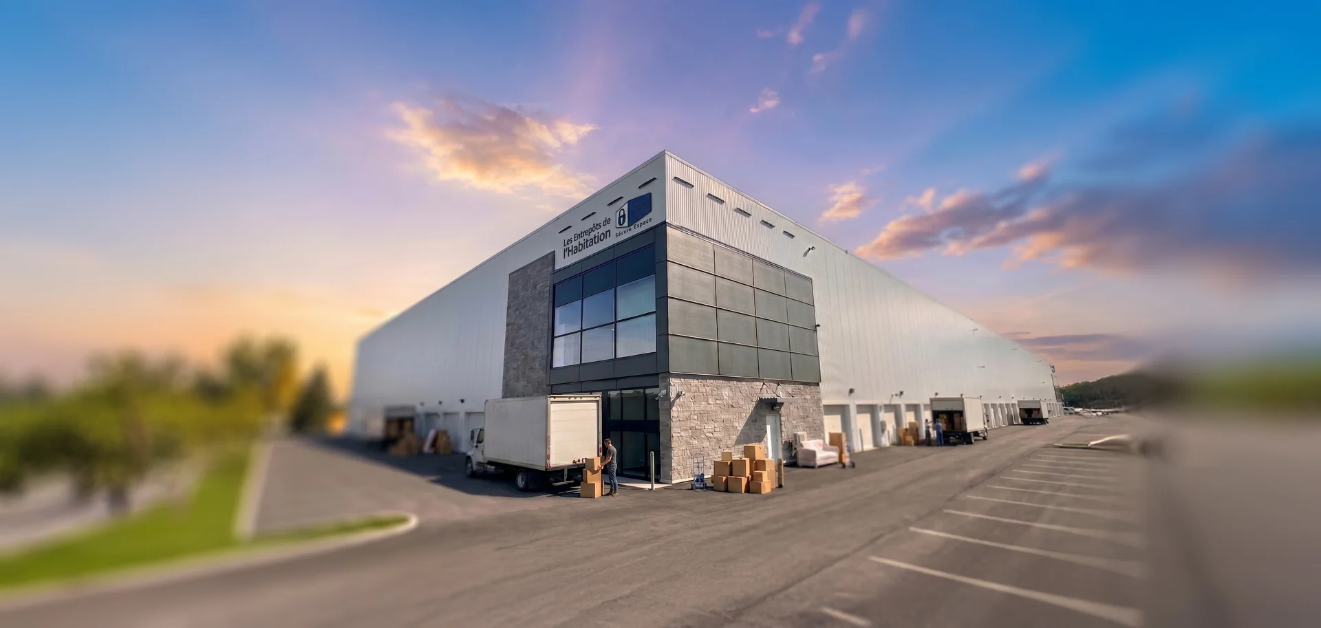 Vue extérieure d’un vaste centre d’entreposage avec plusieurs portes de garage, un camion de déménagement et des boîtes empilées près de l’entrée principale.