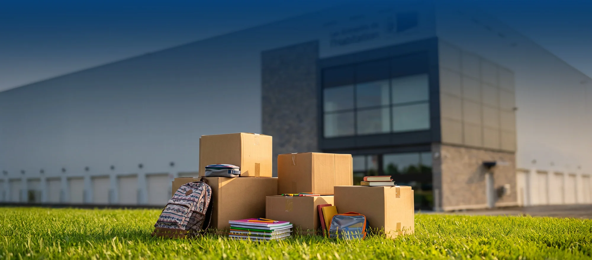 Cartons de déménagement, fournitures scolaires et sac à dos devant un entrepôt moderne, illustrant un service d'entreposage ou de déménagement.